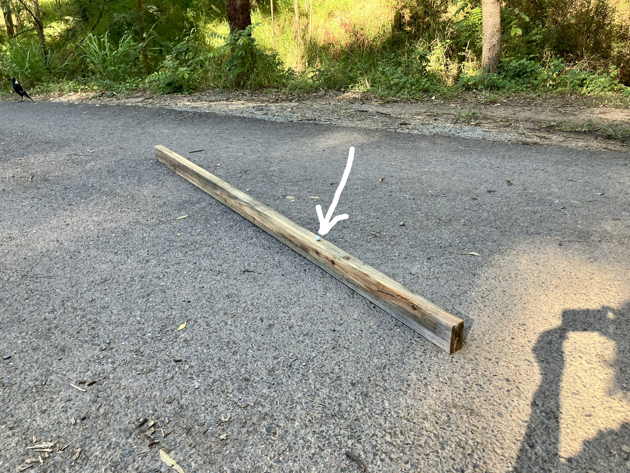 A piece of timber laid out on a bitumen road. It's 45mm by 90mm by 2400mm. There's a white arrow pointing to a white marble resting on top. The marble is about one quarter to one third along the length.